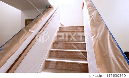A staircase in a home undergoing renovation, featuring protective coverings and masking tape to safeguard surfaces. The area is prepared for painting or further construction work, highlighting a A staircase in a home undergoing renovation, featuring protective coverings and masking tape to safeguard surfaces. The area is prepared for painting or further construction work, highlighting a 115630560