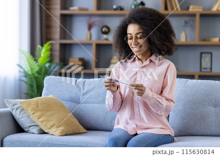 Happy woman holding a positive pregnancy test while sitting on a couch in a comfortable and cozy living room. Happy woman holding a positive pregnancy test while sitting on a couch in a comfortable and cozy living room. 115630814