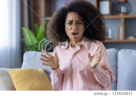 Surprised woman with curly hair and glasses reacting with open hands while sitting on a couch at home Surprised woman with curly hair and glasses reacting with open hands while sitting on a couch at home 115630831