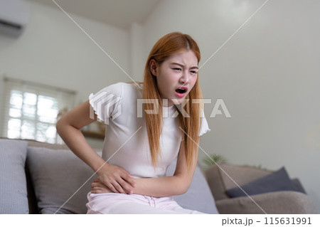 Woman Experiencing Stomach Pain While Sitting on Couch, Holding Her Abdomen in Discomfort 115631991