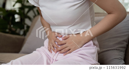 Woman Experiencing Stomach Pain While Sitting on a Couch in a Home Setting, Clutching Her Abdomen in Discomfort 115631992