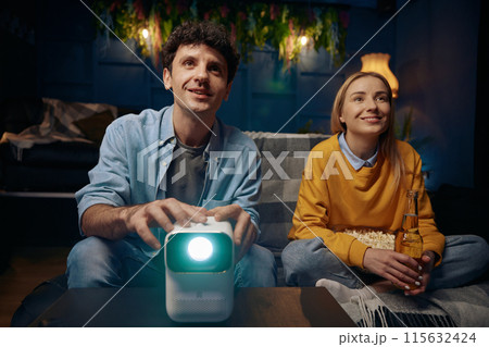 Man and a woman are sitting on a couch watching a movie on a projector 115632424