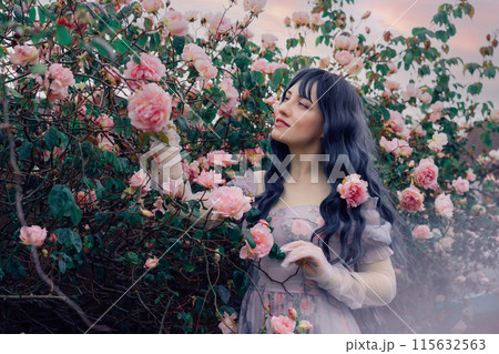 Fairy tale woman portrait. Young beautiful woman Fairy holds pink flower rose on blooming bush background. Hairstyle loose green hair, romantic purple medieval vintage dress. Summer fantasy garden. 115632563