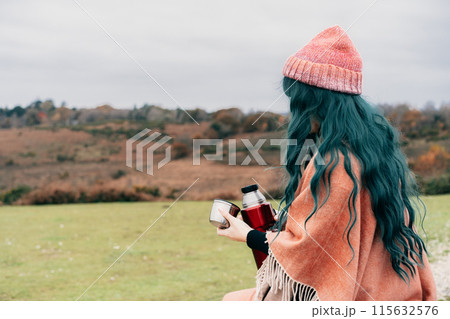 Hipster Woman sits on car's hood, wrapped in warm Shawl Wraps Poncho Cape and hat and enjoying warm drink, while overlooking rural landscape during road trip. Cold season travel, vacation concept Hipster Woman sits on car's hood, wrapped in warm Shawl Wraps Poncho Cape and hat and enjoying warm drink, while overlooking rural landscape during road trip. Cold season travel, vacation concept 115632576