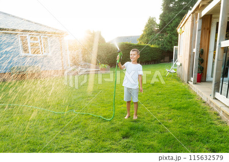 Joyful smiling boy playing with water sprinkler hose, watering grass. Summer garden outdoor fun for children. Boy splashing water on hot sunny day in backyard. Happy active childhood. Selective focus. 115632579