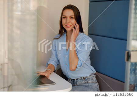 A smiling woman is having a phone call while working on a laptop in a modern office space 115633560