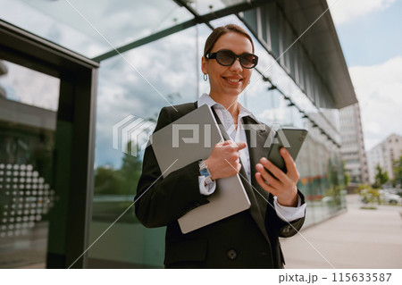Businesswoman using smartphone and carrying laptop outside modern office building. Businesswoman using smartphone and carrying laptop outside modern office building. 115633587
