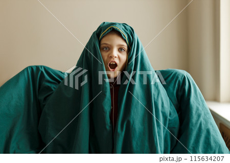 Cheerful little funny girl of 7 wrapped up in green blanket depicts ghost at home 115634207