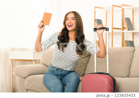 A smiling young woman holds up her passport and boarding passes excitedly as she sits on a wheeled suitcase, prepared to embark on a journey 115635433