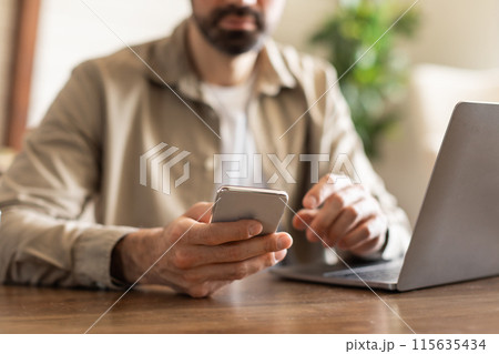 Cropped of man is sitting at a table while actively using a cell phone. He appears engaged with the device, possibly texting, browsing, or talking on the phone. 115635434