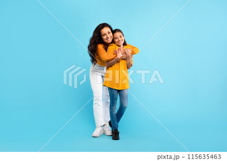 Happy european mother hugging her lovely daughter, standing over blue studio background, full length shot, free space. Young mom and he child girl bonding and smiling to camera 115635463