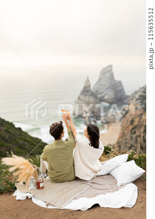 Back view of romantic couple celebrating their engagement or honeymoon on the coastline, drinking champagne and clinking glasses, enjoying ocean shore, copy space, vertical shot Back view of romantic couple celebrating their engagement or honeymoon on the coastline, drinking champagne and clinking glasses, enjoying ocean shore, copy space, vertical shot 115635512