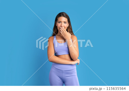 Glad shocked slim young caucasian lady in sportswear covers open mouth with hand after workout, isolated on blue studio background. Surprise emotions, good news, fitness, body care and weight loss 115635564
