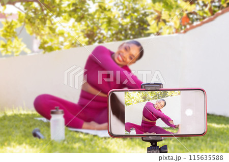 Smartphone screen recording cheerful attractive sporty black woman fitness blogger exercising outdoor at garden. Athletic african american lady fitness coach streaming while stretching body 115635588