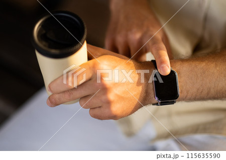 Close up shot of a businessman hand that uses a smart watch to view incoming messages and calls while sitting outdoors and drinking takeaway coffee, the gadget simplifies his life, cropped 115635590