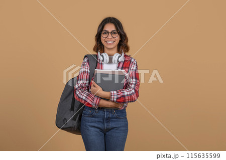 Young middle eastern lady student in a red and white plaid shirt, holding a stack of books and a backpack. She is wearing glasses and smiling at the camera. The background is a plain tan color. Young middle eastern lady student in a red and white plaid shirt, holding a stack of books and a backpack. She is wearing glasses and smiling at the camera. The background is a plain tan color. 115635599