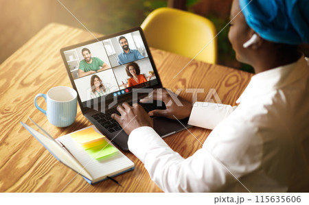 African American woman is sitting at a wooden table, engaging in a virtual meeting on her laptop with four colleagues. She has a colorful book and a mug beside her. 115635606