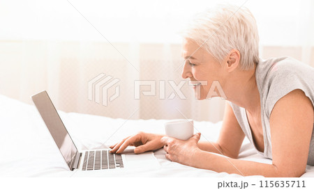 Senior woman is laying on a bed while using a laptop. She appears focused on the screen, with her hands typing or scrolling. The room is dimly lit, creating a cozy atmosphere, side view Senior woman is laying on a bed while using a laptop. She appears focused on the screen, with her hands typing or scrolling. The room is dimly lit, creating a cozy atmosphere, side view 115635711