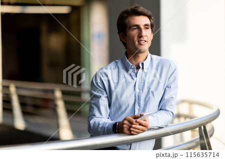 Communication in business concept. Handsome millennial man entrepreneur with phone in his hands standing outdoor by office buildng, businessman waiting for important call, copy space 115635714