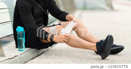 Cropped of runner suffering with knee pain during workout outdoor. Sportsman sitting on footbridge, putting bandage on his leg, panorama with copy space. Exercise and injury prevention concept Cropped of runner suffering with knee pain during workout outdoor. Sportsman sitting on footbridge, putting bandage on his leg, panorama with copy space. Exercise and injury prevention concept 115635778