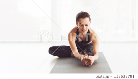 A young woman is practicing a yoga stretch indoors on a mat during the morning. She is wearing comfortable workout attire and appears focused and calm. 115635791