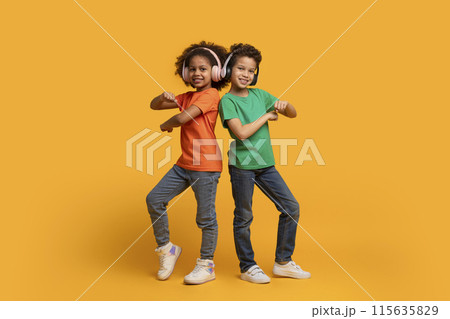 Two African American children with headphones, a boy and a girl, joyfully dancing on a bright yellow background. Their movements are energetic and synchronized 115635829