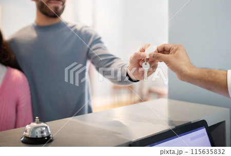 Unrecognizable Travelers Couple Checking In At Hotel Taking Accommodation Room Key From Receptionist Man, Standing At Reception Counter Indoor. Cropped Shot, Selective Focus 115635832