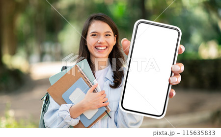 A young woman, likely a student, smiles brightly as she holds a smartphone with a blank screen. She is outdoors in a park-like setting, and her hand is outstretched, presenting the phone to the viewer A young woman, likely a student, smiles brightly as she holds a smartphone with a blank screen. She is outdoors in a park-like setting, and her hand is outstretched, presenting the phone to the viewer 115635833