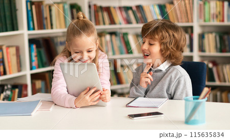 Two primary school kids sitting together at desk in classroom or library interior, using digital tablet computer, playing games or watching educational videos during break 115635834