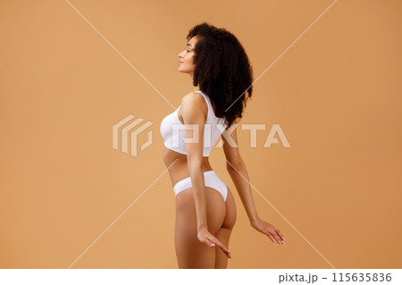 Young slim black lady with perfect ideal body shape posing isolated on beige studio background, half-turned sexy slender woman in white underwear, free space Young slim black lady with perfect ideal body shape posing isolated on beige studio background, half-turned sexy slender woman in white underwear, free space 115635836
