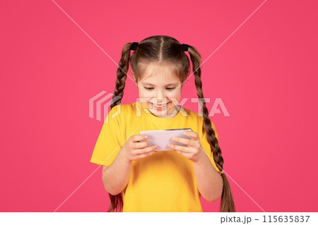 Cheerful preteen girl playing games on smartphone while standing on pink studio background, cute female child looking at mobile phone screen and smiling, enjoying online gaming, copy space 115635837