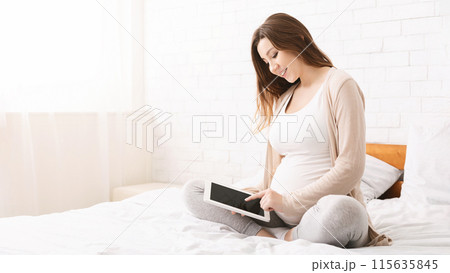 A pregnant woman is sitting cross-legged on a bed and smiling while using a tablet, creating a peaceful atmosphere as she enjoys her time connected to her device, copy space A pregnant woman is sitting cross-legged on a bed and smiling while using a tablet, creating a peaceful atmosphere as she enjoys her time connected to her device, copy space 115635845