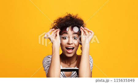 A joyful young African American woman with voluminous curly hair lifts her eyeglasses in a gesture of astonishment, showcasing a beaming smile against a vivid yellow backdrop A joyful young African American woman with voluminous curly hair lifts her eyeglasses in a gesture of astonishment, showcasing a beaming smile against a vivid yellow backdrop 115635849