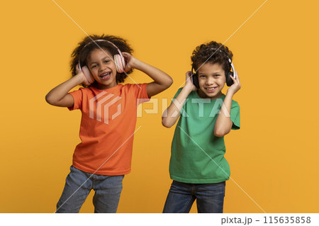 Two African American children stand in front of a yellow background while listening to music with headphones 115635858