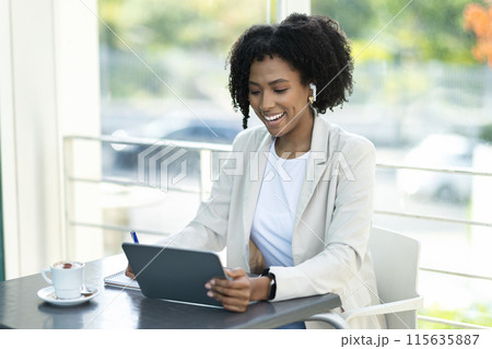 Cheerful attractive millennial black woman in formal outfit sitting at table at terrace, drinking coffee, have video call with business partner, using digital tablet, cafe interior, copy space Cheerful attractive millennial black woman in formal outfit sitting at table at terrace, drinking coffee, have video call with business partner, using digital tablet, cafe interior, copy space 115635887