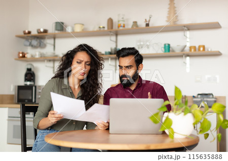 Bankruptcy concept. Overwhelmed arab couple sitting at kitchen desk in front of laptop, holding documents and reading correspondence, got mail from bank 115635888