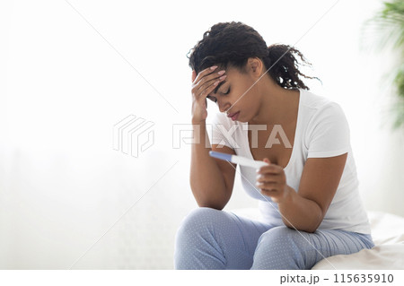 Sad young black woman checking her recent pregnancy test while sitting on bed at home, upset african american lady facing unwanted childbearing or being unable to conceive baby, having health problem 115635910