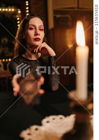Vertical portrait of mysterious fortuneteller asking client to move deck of cards for layout during session, in dark esoteric room by light of burning candle, looking at camera with serious expression Vertical portrait of mysterious fortuneteller asking client to move deck of cards for layout during session, in dark esoteric room by light of burning candle, looking at camera with serious expression 115635918