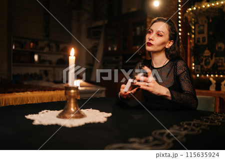 Confident mysterious fortuneteller shuffling deck of tarot cards by light of burning candles, looking away with serious expression. Sorceress shuffles Tarot cards before forecasting future for client Confident mysterious fortuneteller shuffling deck of tarot cards by light of burning candles, looking away with serious expression. Sorceress shuffles Tarot cards before forecasting future for client 115635924