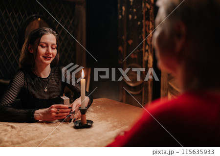 Portrait of fortune teller making tarot layout on table to mature adult female client when predicting future in dark room by light of burning candle. Concept of prophecy, prediction and esoteric. 115635933