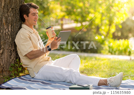 Happy handsome guy wearing casual outfit sitting under tree at public park, using digital tablet, copy space. Millennial man resting outdoors, have video call with family or lover. Telecommunication 115635980