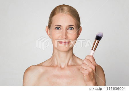A woman with blonde hair and fair skin is shown holding a makeup brush in front of a white background. She is looking directly at the camera and smiling slightly. A woman with blonde hair and fair skin is shown holding a makeup brush in front of a white background. She is looking directly at the camera and smiling slightly. 115636004