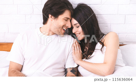 A man and a woman are sitting together in bed, smiling at each other. The man is wearing a white t-shirt and the woman is wearing a white tank top. They look happy and in love. 115636064