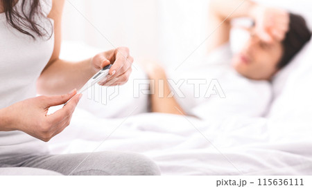 A woman holds a digital thermometer in her hands, while a man lays in bed, sick, and appears to be sleeping. A woman holds a digital thermometer in her hands, while a man lays in bed, sick, and appears to be sleeping. 115636111