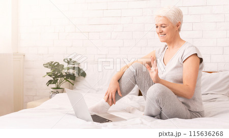 A senior woman is sitting on a bed, intensely focused on using a laptop computer. She appears to be working or engaging in online activities, have video conference 115636268
