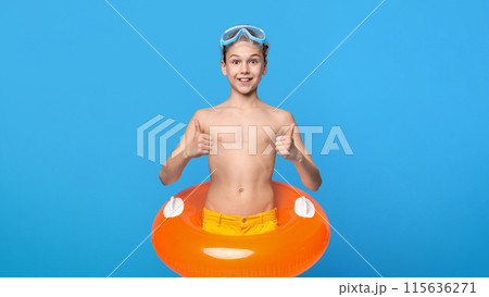 A cheerful boy wearing yellow swim trunks and blue goggles gives a double thumbs-up while standing inside an orange pool float against a bright blue backdrop 115636271