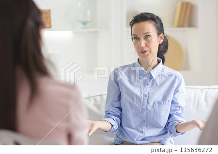 A woman in a light blue shirt is engaging in a counseling session during daytime in a cozy, well-lit office with white walls and minimal decor. She appears attentive and expressive 115636272