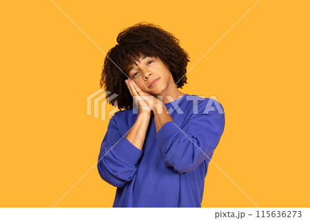 A young man with curly hair is wearing a blue sweater and posing as if he is sleeping, with hands together under his cheek. He stands in front of a vibrant orange background 115636273