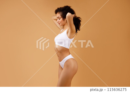 Young slender lady posing in white underwear showing her perfect fit body, holding hands behind her head, standing posing over beige studio background, free space 115636275
