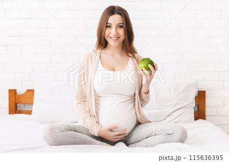 A pregnant woman sitting on a bed, holding a green apple in her hands. She appears calm and content as she gazes at the apple in her hands. A pregnant woman sitting on a bed, holding a green apple in her hands. She appears calm and content as she gazes at the apple in her hands. 115636395